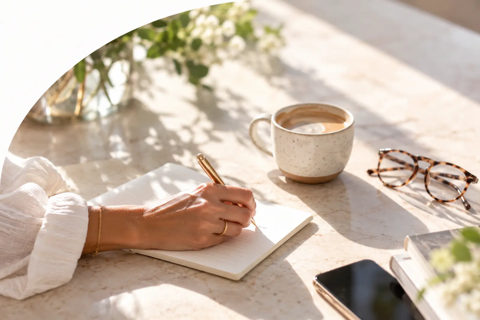 A hand writes in a notebook beside a cup of coffee and reading glasses.